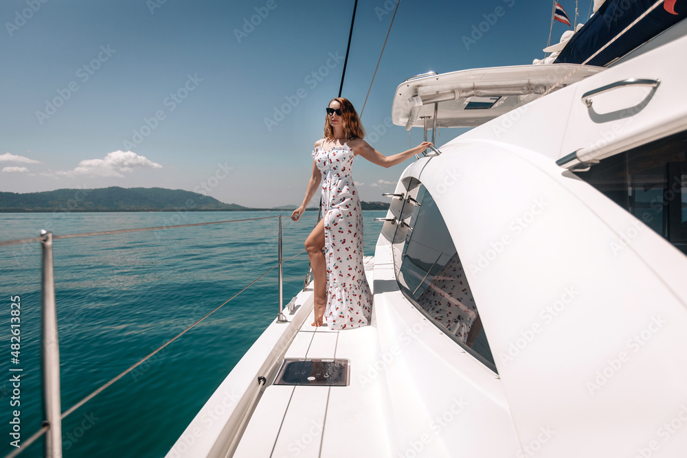 Sexual adorable young lady wearing a long dress and sunglasses and posing on her white yacht against the backdrop of the sea and nature. Summertime concept - obrazy, fototapety, plakaty 