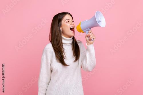 Side view portrait of pretty positive girl screaming in megaphone, announcing important information, wearing white casual style sweater. Indoor studio shot isolated on pink background. photo