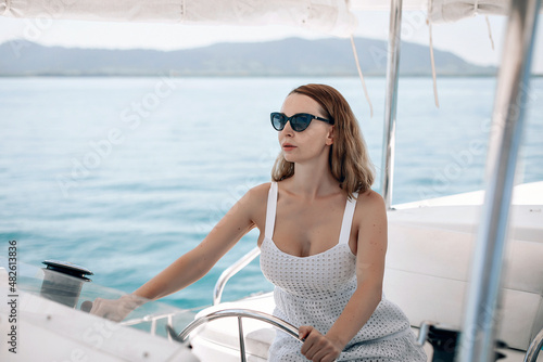 Portrait of a beautiful lovely adult female model in a white dress with an open neckline and sunglasses sitting at the wheel of her yacht and watching the road 