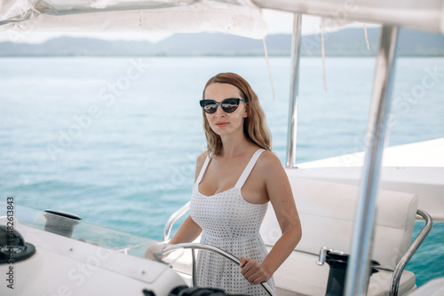 Portrait of a beautiful lovely sexual female model wearing a white dress and sunglasses and sitting at the helm of a yacht, smiling sweetly against the backdrop of another yacht and the sea.