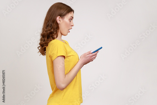 Side view of surprised teenager girl in yellow casual T-shirt, expressing shock while using cell phone, searching web, astonishing news. Indoor studio shot isolated on gray background.