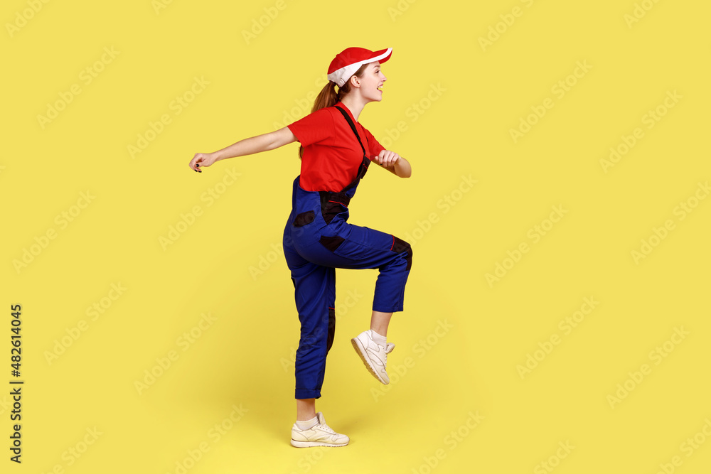 Side view portrait of worker woman marching, going for work, expressing positive emotions and happiness, wearing overalls and red cap. Indoor studio shot isolated on yellow background.