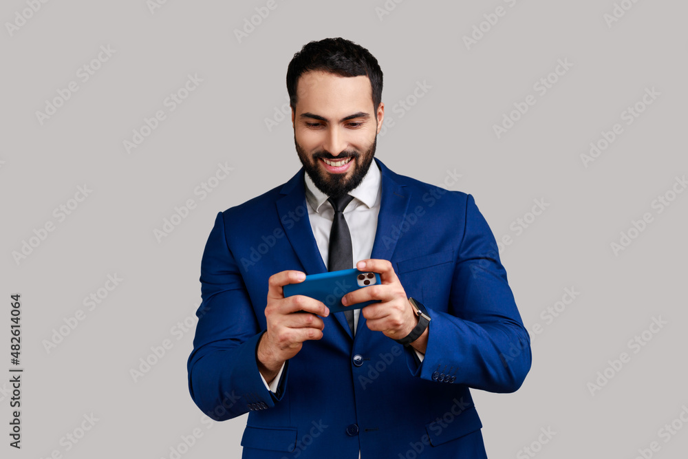 Delighted bearded man using mobile phone with happy positive expression, playing video game on cellphone, wearing official style suit. Indoor studio shot isolated on gray background. Delighted bearded man using mobile phone with happy positive expression, playing video game on cellphone, wearing official style suit. Indoor studio shot isolated on gray background.