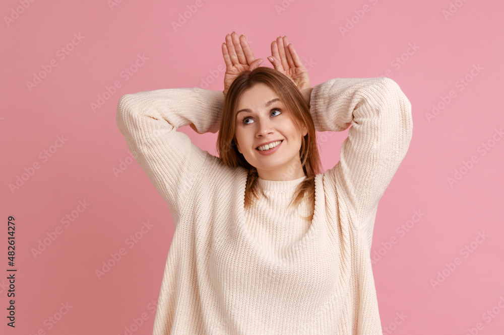 Portrait of blond woman making funny bunny ears with hands on head, childish behavior, playful optimistic mood, wearing white sweater. Indoor studio shot isolated on pink background. Portrait of blond woman making funny bunny ears with hands on head, childish behavior, playful optimistic mood, wearing white sweater. Indoor studio shot isolated on pink background.