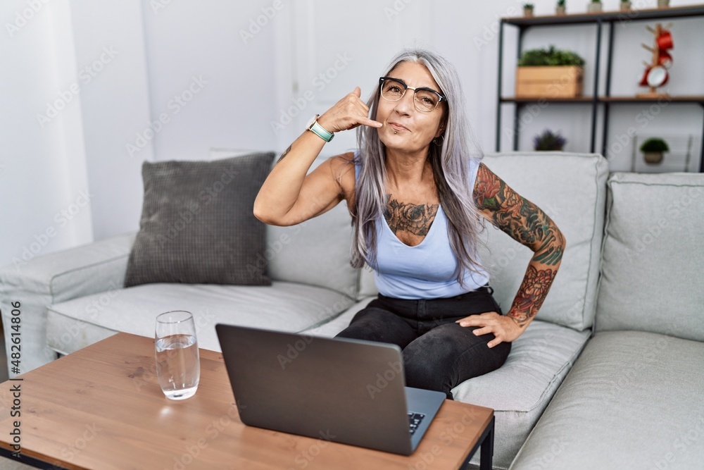 Middle age grey-haired woman using laptop at home smiling doing phone gesture with hand and fingers like talking on the telephone. communicating concepts. Middle age grey-haired woman using laptop at home smiling doing phone gesture with hand and fingers like talking on the telephone. communicating concepts.