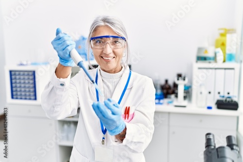 Middle age grey-haired woman wearing scientist uniform using pipette at laboratory