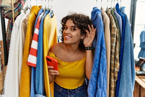 Young hispanic woman searching clothes on clothing rack using smartphone smiling with hand over ear listening an hearing to rumor or gossip. deafness concept.