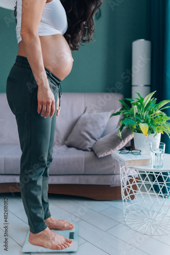 Unrecognisable pregnant barefoot long dark-haired woman staying on scales at home near comfort sofa in modern living room with plant. Female controlling weight during pregnancy. Side view. photo
