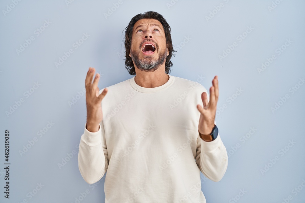 Handsome middle age man wearing casual sweater over blue background crazy and mad shouting and yelling with aggressive expression and arms raised. frustration concept. Handsome middle age man wearing casual sweater over blue background crazy and mad shouting and yelling with aggressive expression and arms raised. frustration concept.
