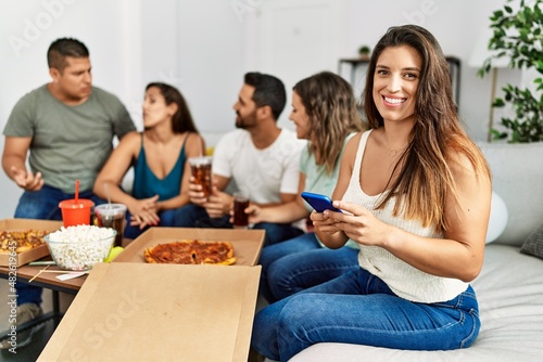 Group of young hispanic friends eating italian pizza sitting on the sofa. Woman smiling happy and using smartphone at home.