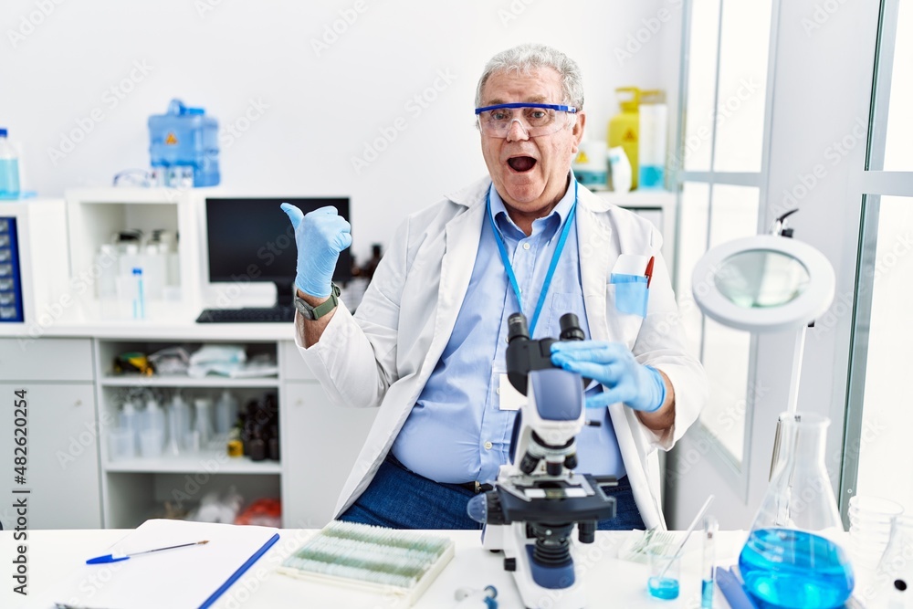 Senior caucasian man working at scientist laboratory surprised pointing with hand finger to the side, open mouth amazed expression. Senior caucasian man working at scientist laboratory surprised pointing with hand finger to the side, open mouth amazed expression.