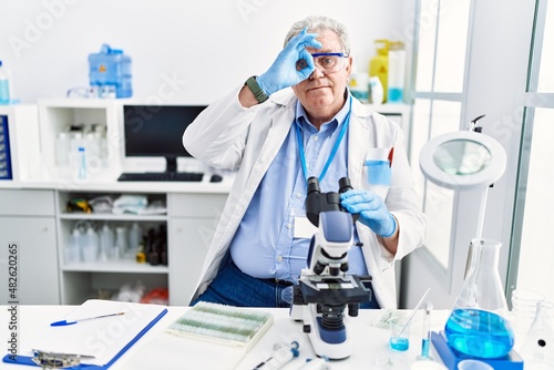 Senior caucasian man working at scientist laboratory doing ok gesture with hand smiling, eye looking through fingers with happy face.