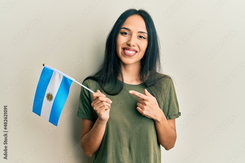 Young hispanic girl holding argentina flag smiling happy pointing with hand and finger - obrazy, fototapety, plakaty 
