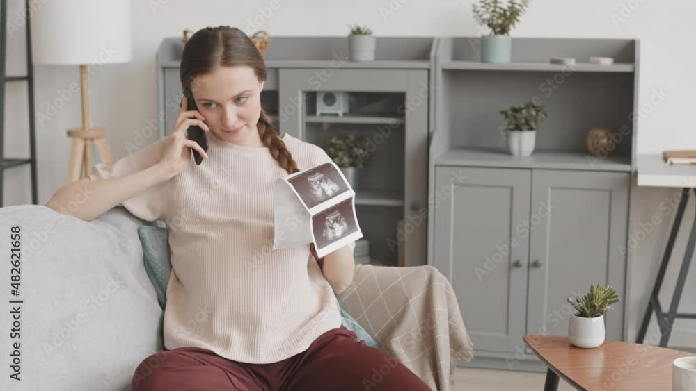 Medium stab shot with slowmo of happy young Caucasian pregnant woman with ultrasound scan in hands sitting on sofa at home while calling father of her baby to bring good news Medium stab shot with slowmo of happy young Caucasian pregnant woman with ultrasound scan in hands sitting on sofa at home while calling father of her baby to bring good news