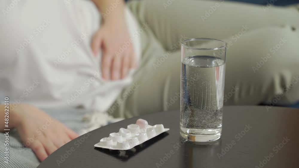 There are glass of water and pills for pain on dark wooden table. Woman lies on sofa and massages stomach with hand from severe pain and cramps. Menstruation, pregnancy, gluttony, pain, cramps There are glass of water and pills for pain on dark wooden table. Woman lies on sofa and massages stomach with hand from severe pain and cramps. Menstruation, pregnancy, gluttony, pain, cramps