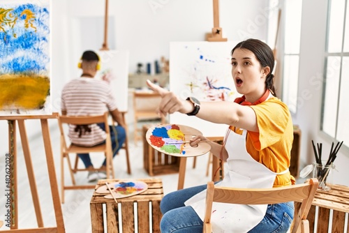 Young artist woman painting on canvas at art studio pointing with finger surprised ahead, open mouth amazed expression, something on the front