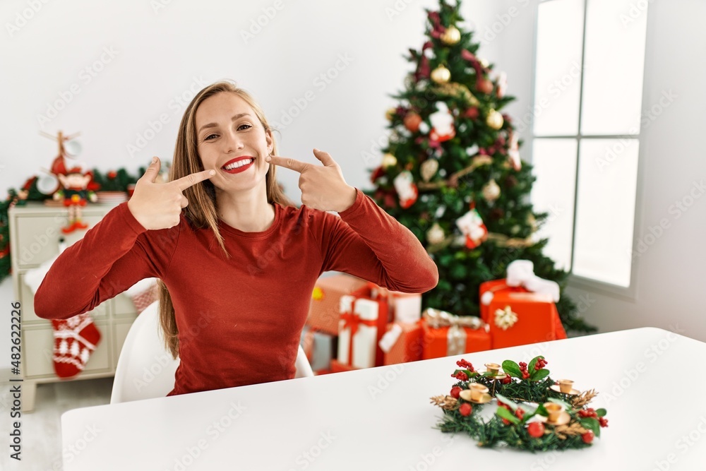 Caucasian young blonde woman sitting on the table by christmas tree smiling cheerful showing and pointing with fingers teeth and mouth. dental health concept. Caucasian young blonde woman sitting on the table by christmas tree smiling cheerful showing and pointing with fingers teeth and mouth. dental health concept.