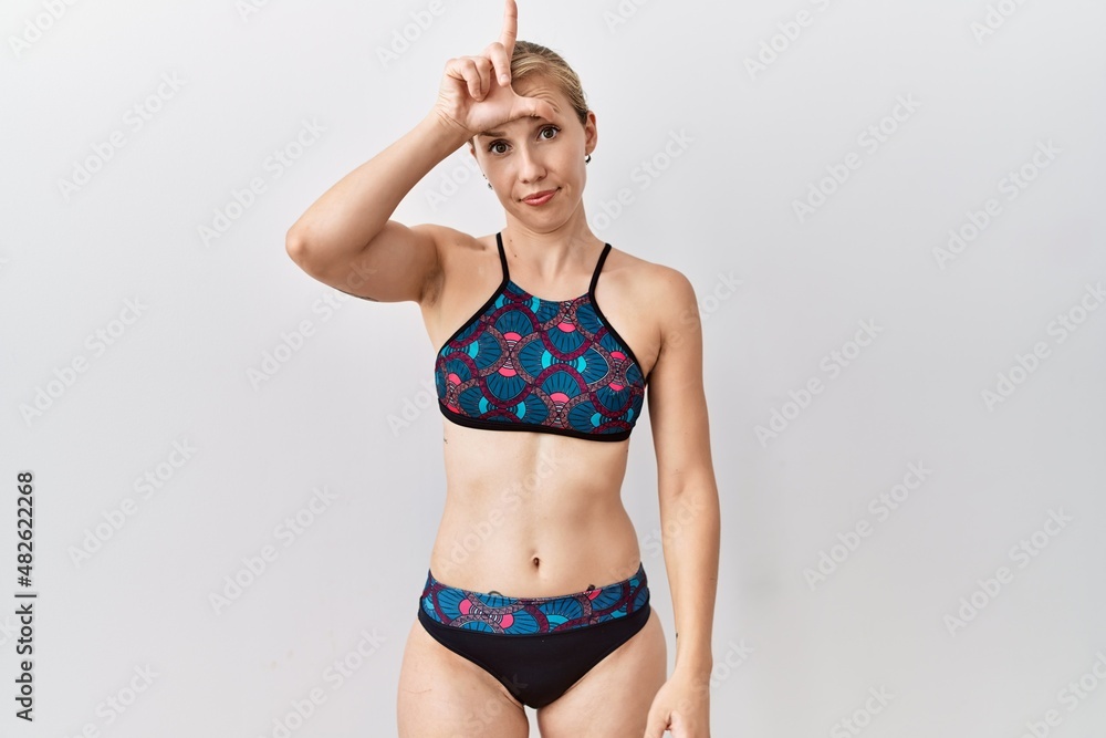 Fototapeta premium Young blonde woman wearing sporty bikini over isolated background making fun of people with fingers on forehead doing loser gesture mocking and insulting.