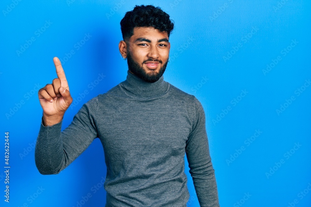 Fototapeta premium Arab man with beard wearing turtleneck sweater pointing finger up with successful idea. exited and happy. number one.