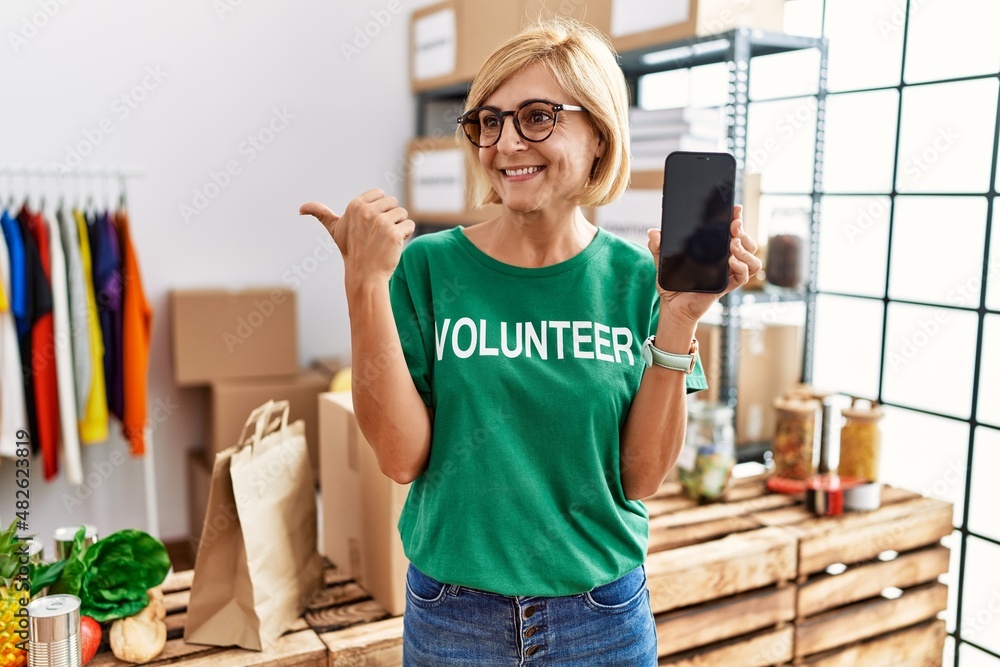 Middle age blonde woman working wearing volunteer t shirt showing smartphone screen pointing thumb up to the side smiling happy with open mouth