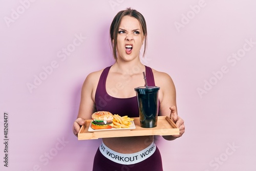 Young hispanic girl wearing sportswear eating a tasty classic burger with fries and soda angry and mad screaming frustrated and furious, shouting with anger. rage and aggressive concept.