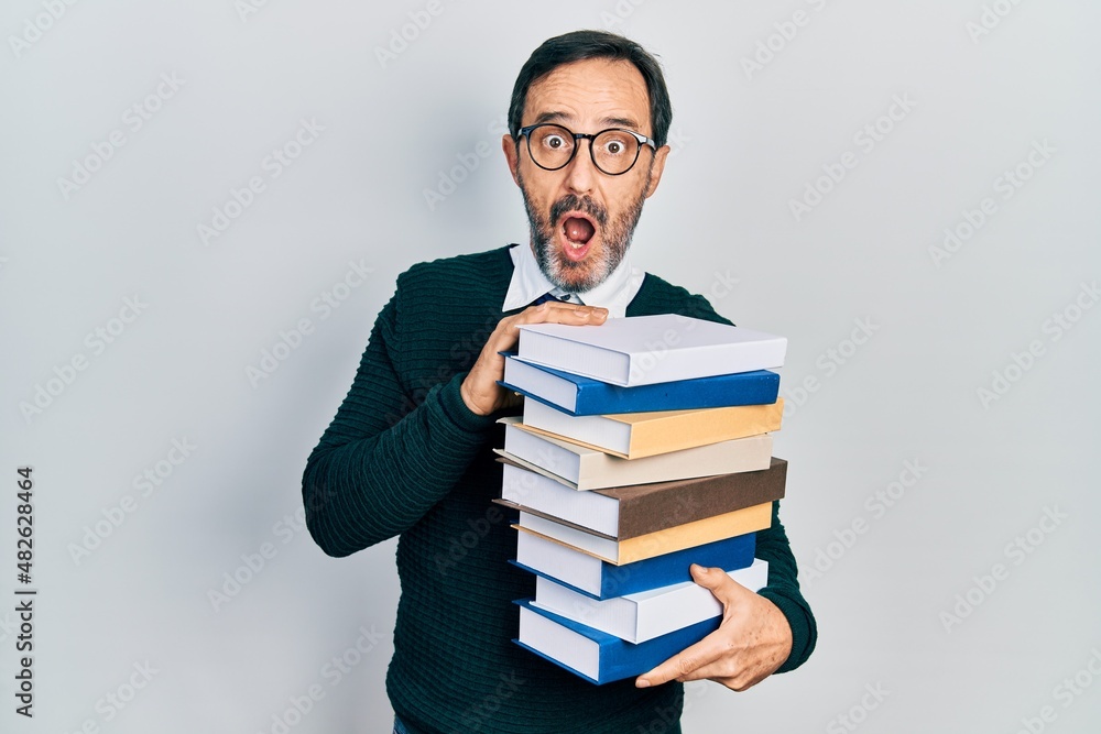 Middle age hispanic man holding a pile of books afraid and shocked with surprise and amazed expression, fear and excited face.