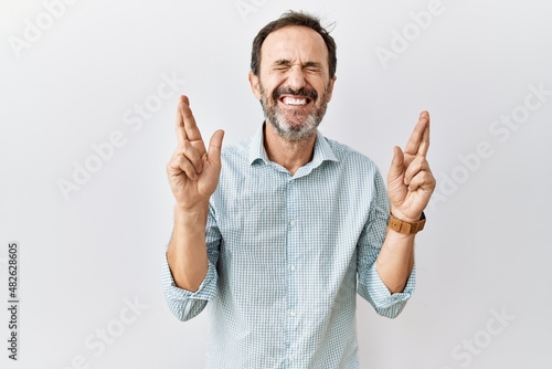 Middle age hispanic man with beard standing over isolated background gesturing finger crossed smiling with hope and eyes closed. luck and superstitious concept. photo