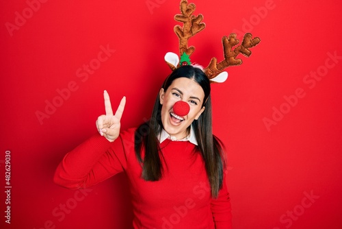 Young hispanic woman wearing deer christmas hat and red nose smiling looking to the camera showing fingers doing victory sign. number two.