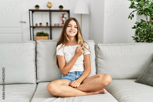 Young brunette teenager sitting on the sofa at home cheerful with a smile of face pointing with hand and finger up to the side with happy and natural expression on face