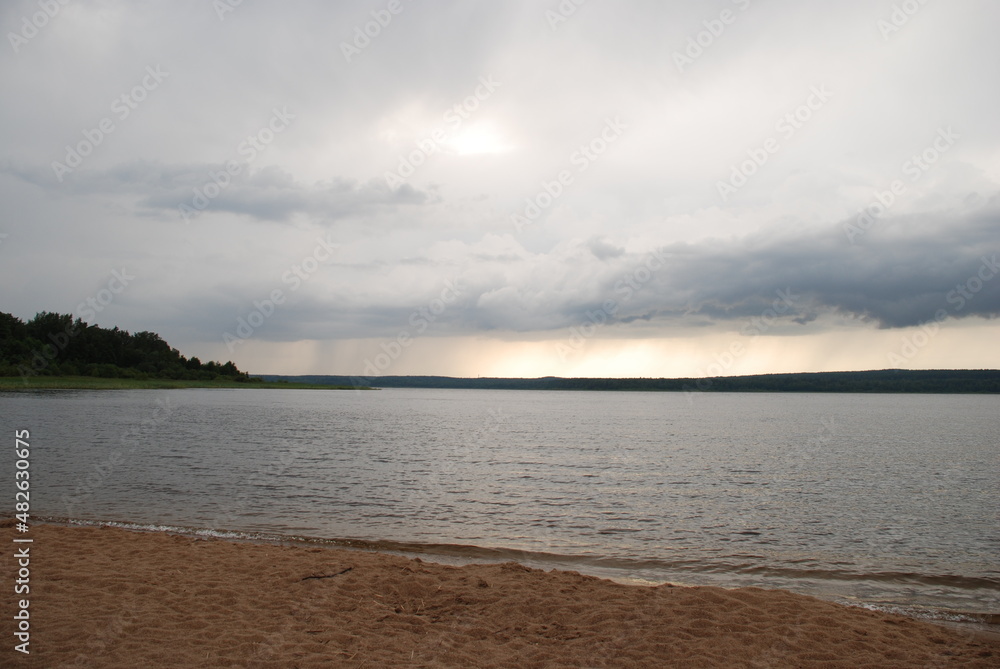 Wide flood of the Vuoksa river. Beach brown sand, river with clear clean water on it small waves. From above, white-gray with blue clouds from which it is raining in the distance. Wide flood of the Vuoksa river. Beach brown sand, river with clear clean water on it small waves. From above, white-gray with blue clouds from which it is raining in the distance.