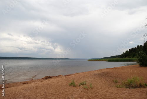 Fototapeta Naklejka Na Ścianę i Meble -  Brown beach sand on the river bank. Trees, bushes and grass grow on the shore. The part is covered with brown sand. The river is wide with clear clear water. Reeds grow in the water.