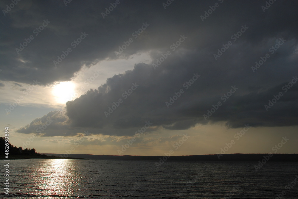 Gray menacing clouds over the river. The clouds are gray-white with a blue tint in the sky. The sun's rays shine through them. Below is a wide river and a bank overgrown with forest. Gray menacing clouds over the river. The clouds are gray-white with a blue tint in the sky. The sun's rays shine through them. Below is a wide river and a bank overgrown with forest.