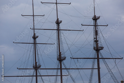 three wooden masts of a Dutch fluyt merchant sailing ship of XVIII century on a grey sky and clouds background close up view
