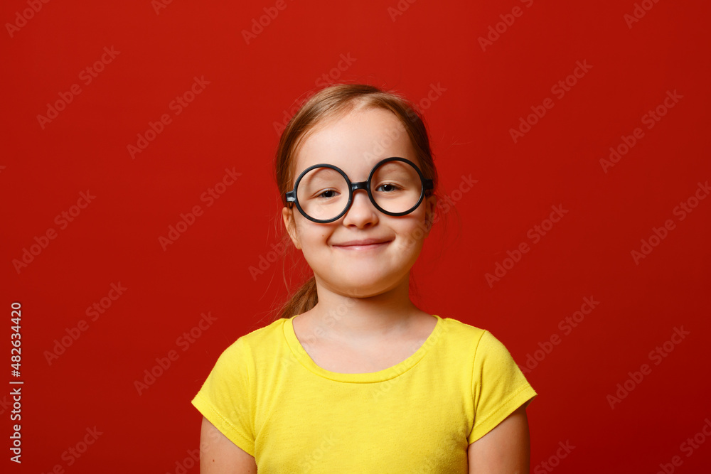 Funny little girl with glasses. Schoolgirl child in a yellow t-shirt on a red isolated background.