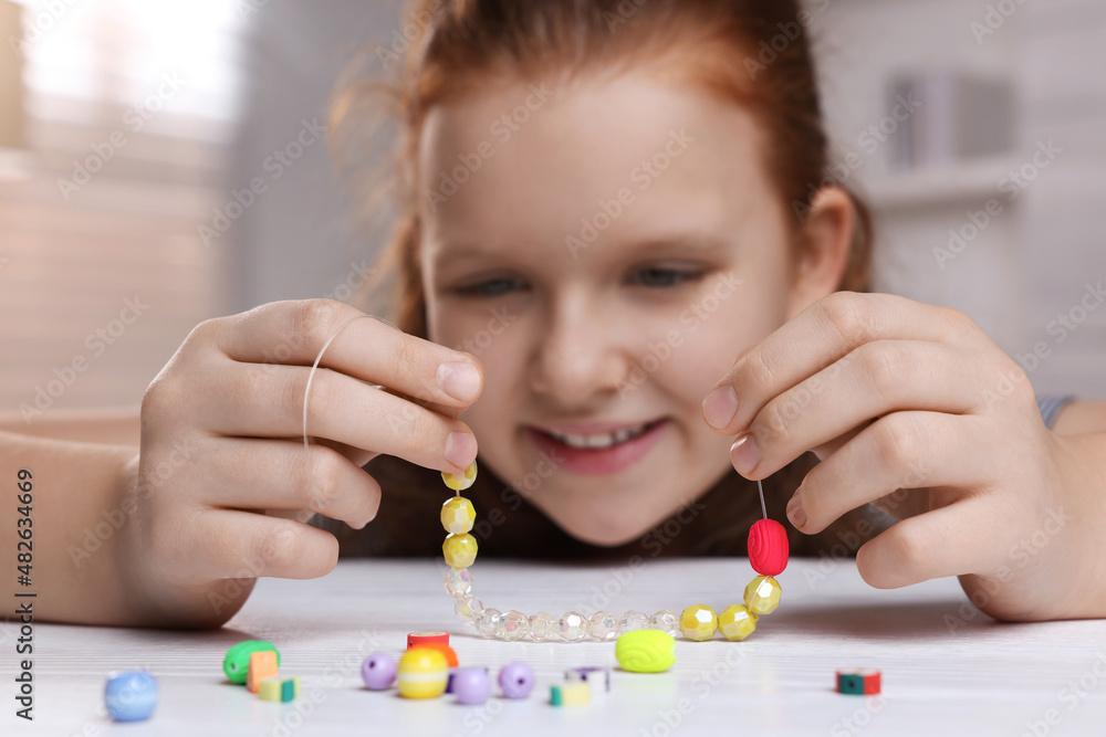 Cute girl making beaded jewelry at table in room, focus on hands Cute girl making beaded jewelry at table in room, focus on hands