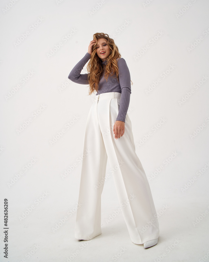 Blonde young woman with stylish hairstyle in knitted sweater and white trousers with high heel shoes clutches head in light studio