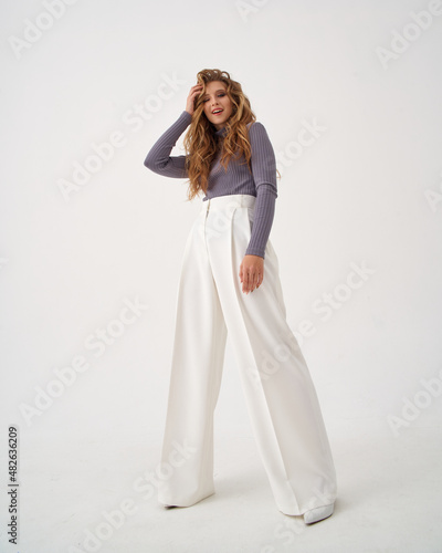 Blonde young woman with stylish hairstyle in knitted sweater and white trousers with high heel shoes clutches head in light studio