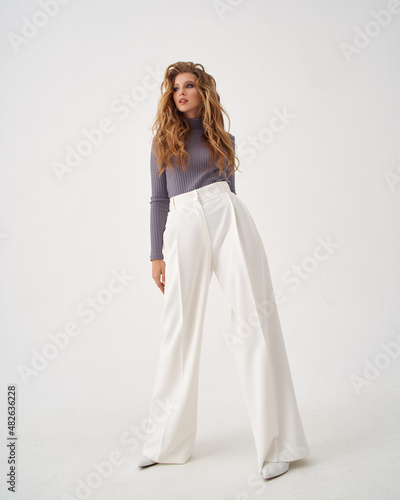 Pretty woman with stylish makeup and long flowing hair wearing sweater and loose trousers holds hand on hip on white background © Dmitry Tsvetkov