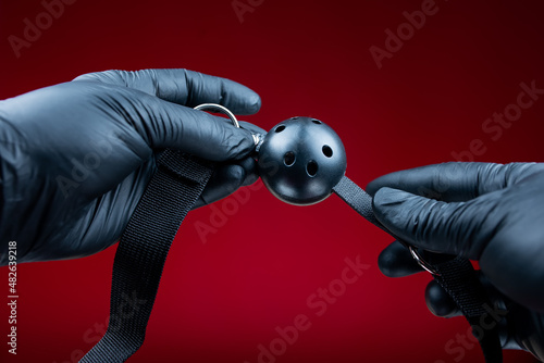 hands in black gloves holding a round gag in the mouth for erotic bdsm games
