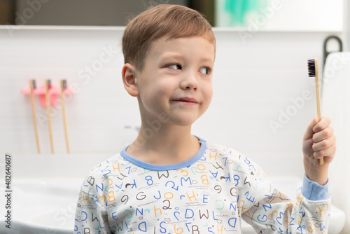 A cute seven-year-old boy in pajamas with a bamboo toothbrush in his hand before going to bed at home in the bathroom against the backdrop of a washbasin with a faucet. Selective focus. Portrait