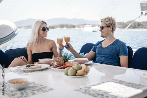 Charming adult couple man and woman are having dinner at the table of their huge and expensive white yacht, smiling and holding glasses of juice in their hands, enjoying each other's company