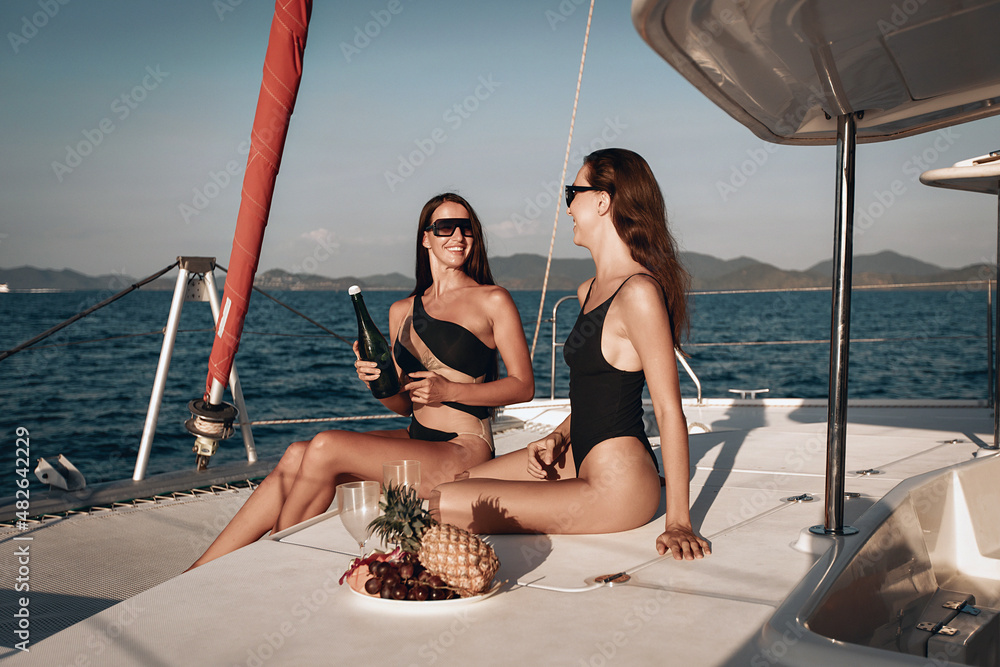 Two cheerful and joyful young girls in black bodysuit swimsuit and sunglasses are happily spending their summer holidays drinking champagne and eating fruit on their huge white yacht. Vacation concept