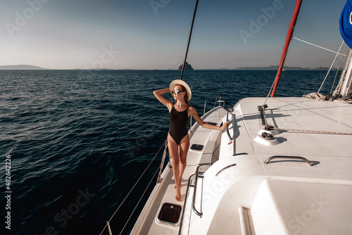 Sexual chining and pretty lady wearing a black tight bikini bodysuit, sunglasses and a panama hat, posing on her laxury white yacht against a backdrop of water and sky. Vacation concept