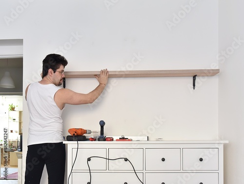Man mounting shelves on the wall. All necessary tools are placed on the chest of drawers