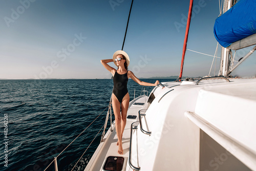 Adorable long-legged young model wearing a black tight bikini bodysuit, sunglasses and a panama hat, posing on her laxury white yacht against a backdrop of water and sky. Holiday concept