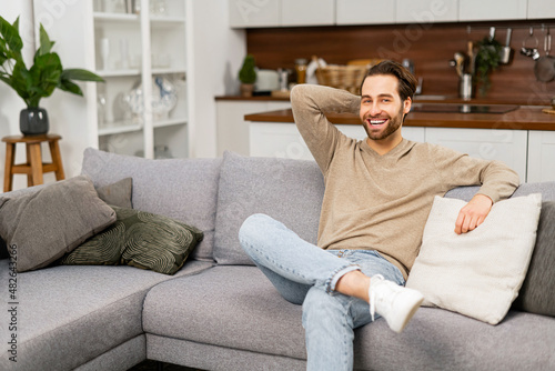 Portrait of young caucasian man resting on sofa, thinking about the future. Handsome positive man with hand on the head sitting in living room and relaxing at home © Vadim Pastuh