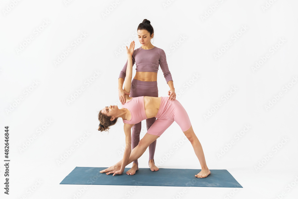 Fototapeta premium A female trainer helps a student to correctly perform the trikonasana exercise, triangle pose, stand on a mat on a white background