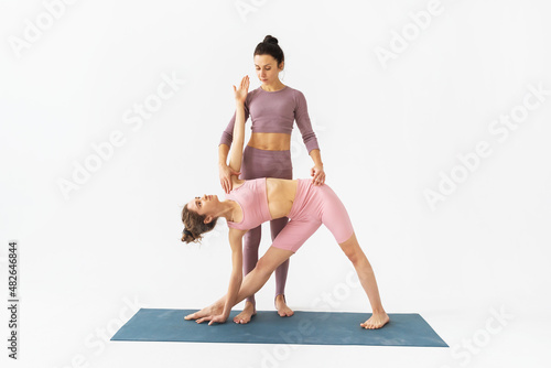 A female trainer helps a student to correctly perform the trikonasana exercise, triangle pose, stand on a mat on a white background © Sergey Chayko