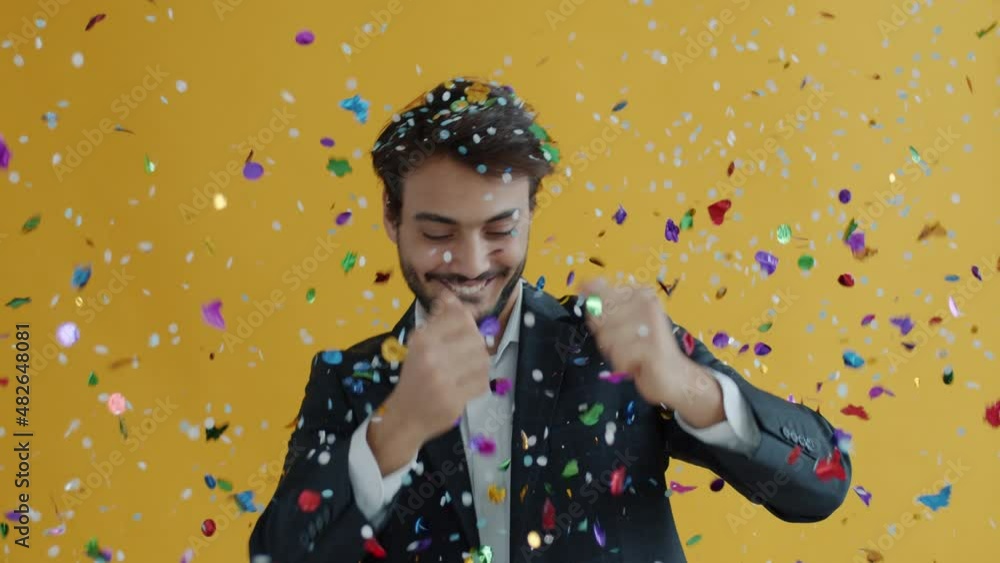 Portrait of Middle Eastern dancer moving while confetti falling down in air during holiday celebration. Man in elegant suit dancing on yellow background. Portrait of Middle Eastern dancer moving while confetti falling down in air during holiday celebration. Man in elegant suit dancing on yellow background.