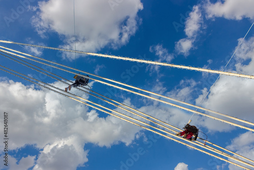 industrial mountaineers repair lighting equipment on a suspension bridge. Construction climbers hang on ropes against blue sky background with clouds